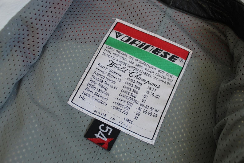 May include: Close-up of a black leather motorcycle jacket interior with a white tag. The tag features the Dainese logo, the words "World Champions," and a list of names and race years. A black tag with the number "54" is also visible.