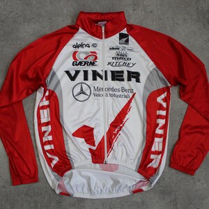 May include: A white and red long-sleeved cycling jersey with the word "VINNER" printed on the front and back. The jersey has a zipper closure and features the logos of several sponsors, including Mercedes-Benz, Gaerne, and Ritchey.