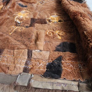 May include: A brown patchwork coat with a fur lining. The coat features various shades of brown, with embroidered floral and scroll designs in gold and white thread. The edges are trimmed with a darker brown material.