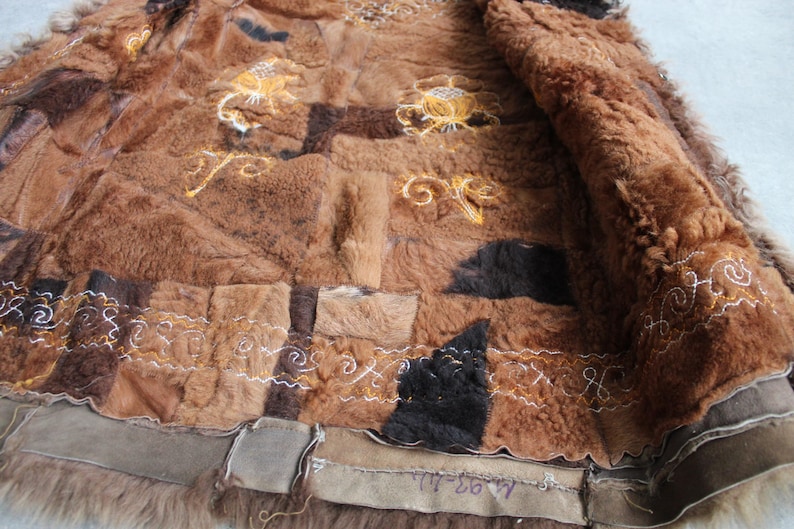 May include: A brown patchwork coat with a fur lining. The coat features various shades of brown, with embroidered floral and scroll designs in gold and white thread. The edges are trimmed with a darker brown material.