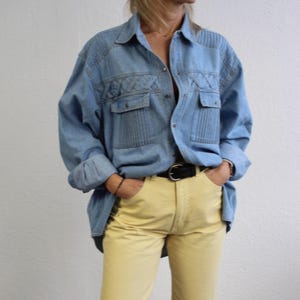 May include: A light blue denim shirt with a decorative stitching pattern on the front. The shirt is unbuttoned and has a black belt around the waist. The person is wearing yellow pants.
