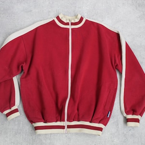 May include: A red zip-up jacket with cream-colored accents. The jacket features a high collar, white stripes down the sleeves, and cream-colored cuffs and hem. It has a full-length zipper and two side pockets. The jacket is made of a soft material.
