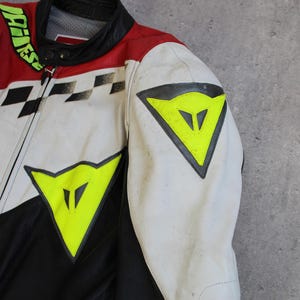 May include: A close-up of a motorcycle racing jacket. The jacket features a color scheme of red, white, and black, with a checkered pattern and neon yellow accents. The jacket has a zipper closure and a black collar.