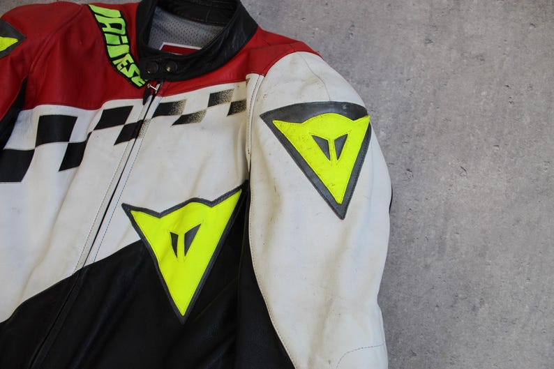 May include: A close-up of a motorcycle racing jacket. The jacket features a color scheme of red, white, and black, with a checkered pattern and neon yellow accents. The jacket has a zipper closure and a black collar.