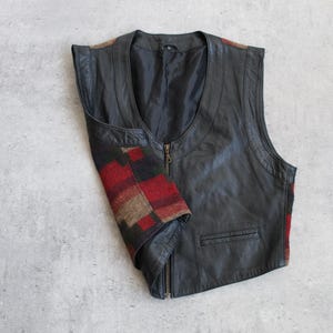 Peut inclure: Un gilet en cuir noir avec une fermeture éclair et une petite poche. Le gilet présente un motif à carreaux rouge, marron et noir sur le côté. Sans manches, il a un col en V.