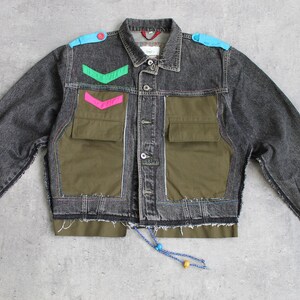 Könnte beinhalten: Graue Jeansjacke mit olivgrünen Taschen und farbigen Akzenten. Die Jacke hat eine Knopfleiste, zwei Brusttaschen und einen Kordelzug am Saum. Dekorative Elemente sind blaue Schulterpolster und grüne und rosa Chevron-Designs.