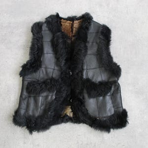 Puede incluir: Chaleco de cuero negro con ribete de piel. El chaleco tiene una parte delantera abotonada y un interior marrón. El ribete de piel está alrededor del cuello, las sisas y el dobladillo inferior.