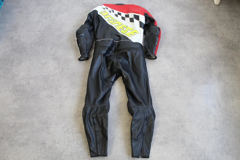 May include: Black and red motorcycle racing suit with white and yellow accents. The suit features a checkered design and the brand name "DAINESE" in yellow. The suit is made of leather and has protective padding on the shoulders and elbows.