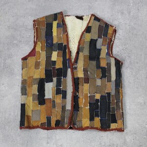May include: A vintage vest with a patchwork design. The vest features a variety of rectangular patches in shades of brown, black, and tan. The interior lining is a cream-colored, fuzzy material. The vest has a V-neck and a button closure.