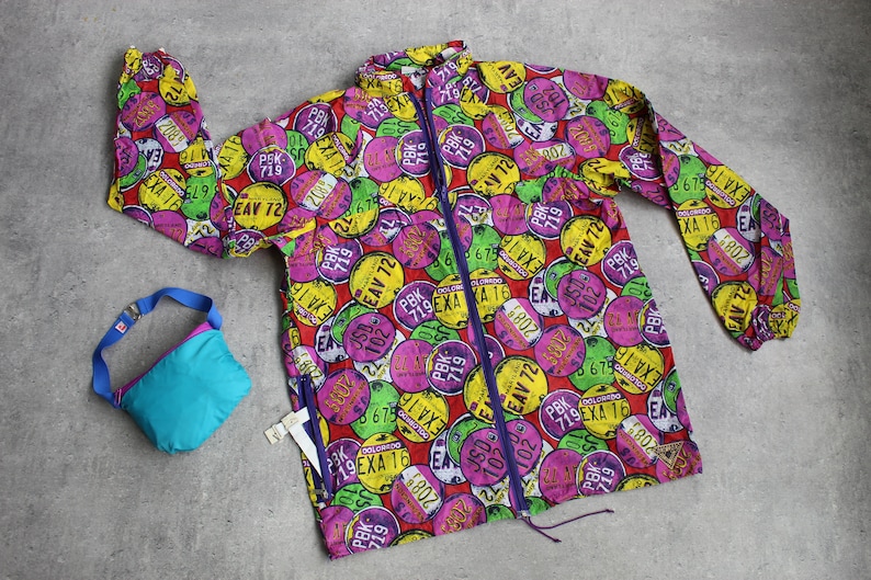 May include: A colorful, lightweight jacket with a zipper closure. The jacket is covered in a pattern of colorful circles with numbers and letters printed on them. The jacket is made of a water-resistant material.