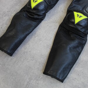 May include: Close-up of black leather motorcycle pants with yellow and white accents on the knee pads. The pants have a textured appearance and are designed for protection. The brand name is visible in gray lettering.