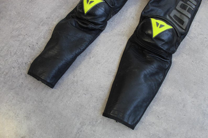 May include: Close-up of black leather motorcycle pants with yellow and white accents on the knee pads. The pants have a textured appearance and are designed for protection. The brand name is visible in gray lettering.