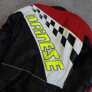 May include: Black and red motorcycle jacket with white and black checkered accents. The jacket features the brand name "DAINESE" in large, neon yellow letters. The jacket appears to be made of leather.