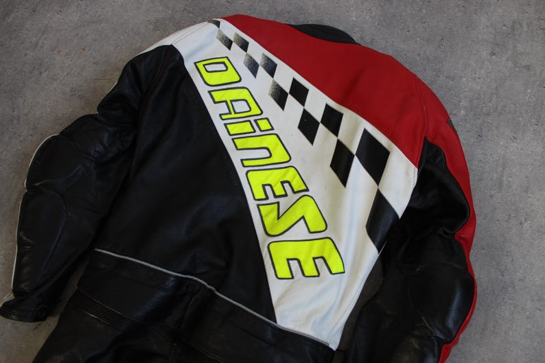 May include: Black and red motorcycle jacket with white and black checkered accents. The jacket features the brand name "DAINESE" in large, neon yellow letters. The jacket appears to be made of leather.