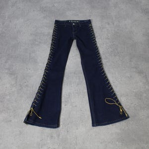May include: Dark blue denim jeans with a flared leg design. The jeans feature gold-colored lacing details along the outer seams. The jeans are laid flat on a gray surface.