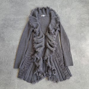 May include: A gray, long-sleeved cardigan with a ruffled trim and a textured knit design. The cardigan features a V-neck and a decorative fur-like trim along the front and collar. The garment is displayed on a gray surface.