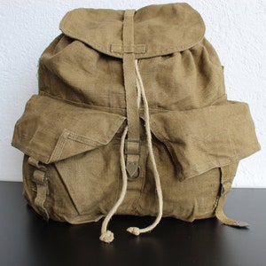 Könnte beinhalten: Ein großer brauner Canvas-Rucksack mit zwei großen Klappentaschen und einem Kordelzugverschluss. Der Rucksack hat einen einzigen Riemen und eine Metallschnalle.