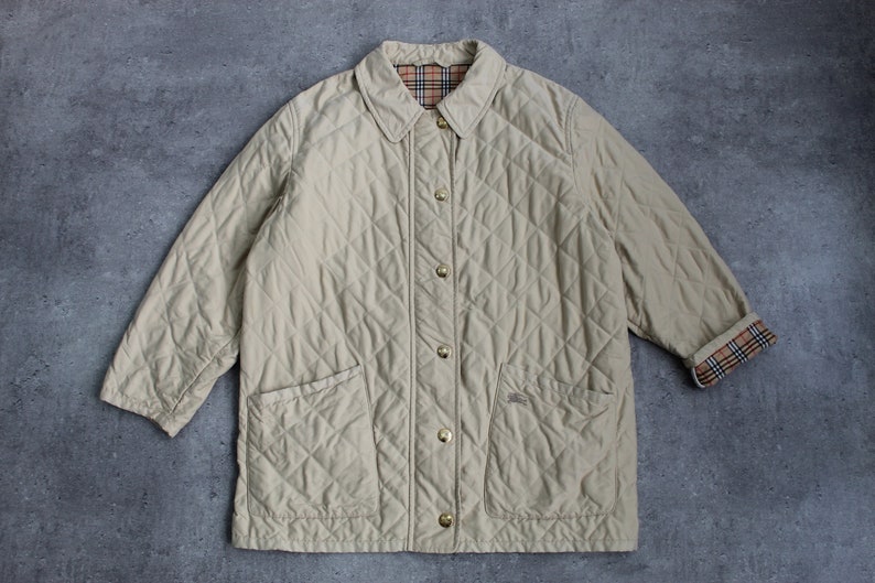 May include: A beige quilted jacket with a button-up front and two pockets. The jacket has a plaid lining visible at the collar and cuffs.