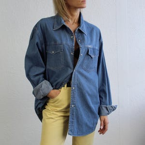 May include: A woman wearing a blue denim shirt with a yellow pair of pants. The shirt has a button-down front and long sleeves.