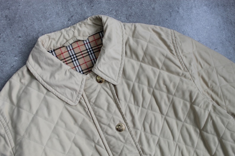 May include: A beige quilted jacket with a plaid lining and gold buttons. The jacket has a classic design and is perfect for layering in cooler weather.