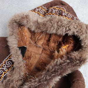 May include: Brown hooded coat with a fur-lined hood and trim. The coat features decorative stitching in white and gold. The interior of the hood is a lighter brown color, and the coat appears to be made of suede or leather.