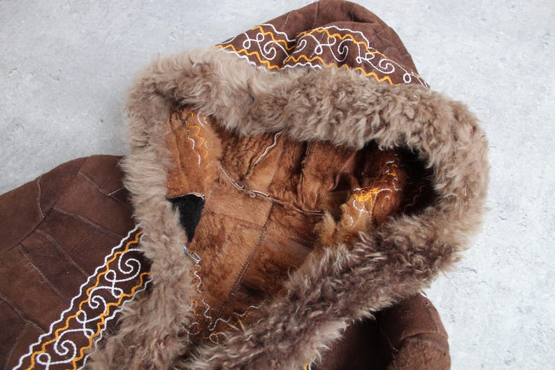 May include: Brown hooded coat with a fur-lined hood and trim. The coat features decorative stitching in white and gold. The interior of the hood is a lighter brown color, and the coat appears to be made of suede or leather.