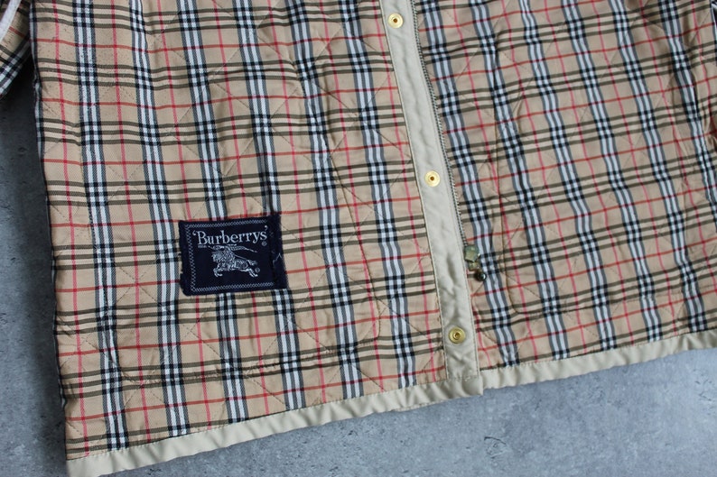 May include: A beige quilted jacket with a black and red plaid pattern. The jacket has a zipper closure and a Burberrys label on the left side.