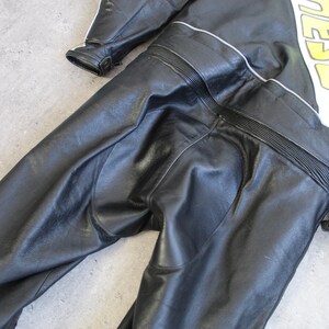 May include: Black leather motorcycle racing suit with white and yellow accents. The suit features a checkered design and the word "DAINESE" in yellow. The suit has a streamlined design with reinforced panels and a red accent.