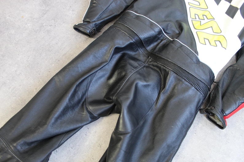 May include: Black leather motorcycle racing suit with white and yellow accents. The suit features a checkered design and the word "DAINESE" in yellow. The suit has a streamlined design with reinforced panels and a red accent.