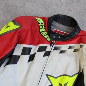 May include: A red, white, and black motorcycle jacket with a black collar. The jacket features a checkered pattern, yellow accents, and the brand name in green. The jacket is made of leather.
