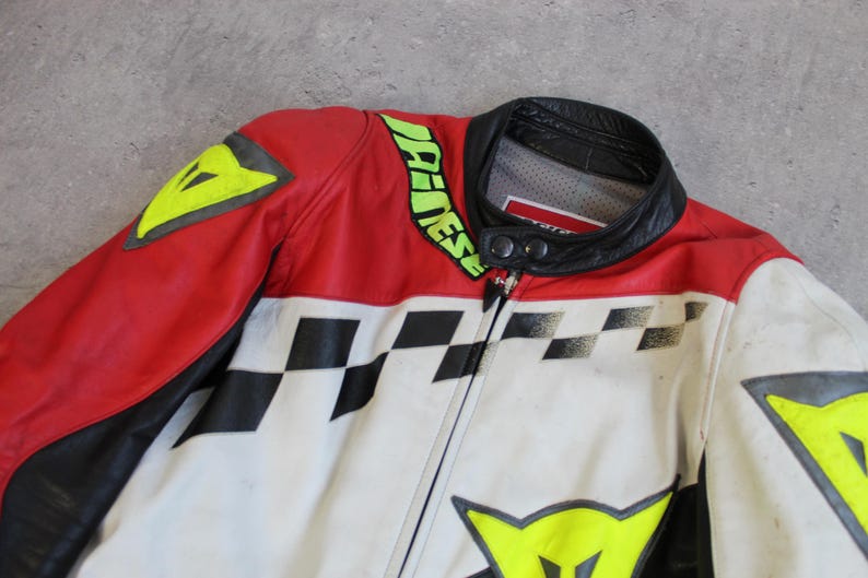 May include: A red, white, and black motorcycle jacket with a black collar. The jacket features a checkered pattern, yellow accents, and the brand name in green. The jacket is made of leather.