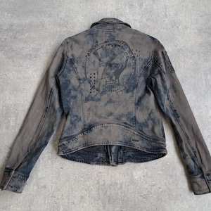 Könnte beinhalten: Grau-blaue Jeansjacke mit verblichenem, abgenutztem Aussehen. Auf der Rückseite befindet sich ein grafisches Design mit Nieten. Die Jacke hat lange Ärmel, einen Knopfverschluss und einen Kragen.