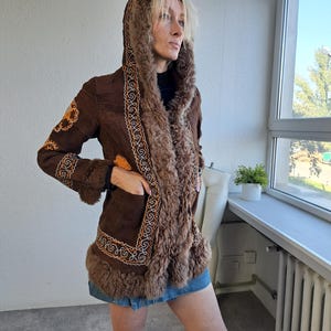 May include: A brown suede coat with a faux fur-lined hood and trim. The coat features intricate embroidered detailing in shades of orange and cream along the edges and sleeves. The coat is worn with a denim skirt and boots.