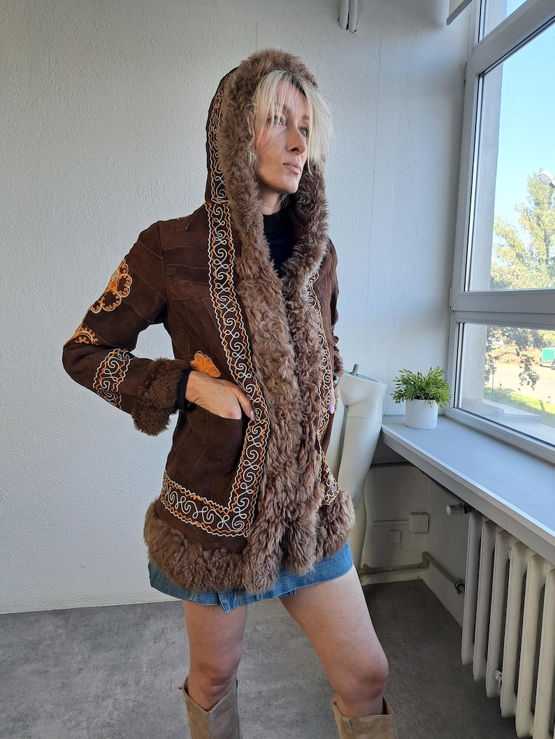 May include: A brown suede coat with a faux fur-lined hood and trim. The coat features intricate embroidered detailing in shades of orange and cream along the edges and sleeves. The coat is worn with a denim skirt and boots.