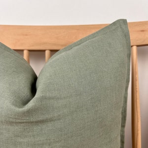 May include: A soft, green linen pillow with a subtle texture. The pillow is placed on a wooden chair with a slatted back.