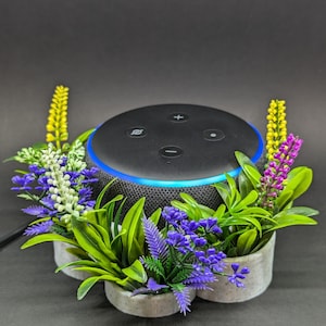Peut inclure: Un haut-parleur intelligent noir avec un anneau bleu autour du haut, entouré de plantes artificielles dans un pot blanc. Le haut-parleur a des boutons pour le volume, la désactivation du microphone et d'autres fonctions.