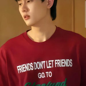 May include: A red crew neck t-shirt with white text that reads "FRIENDS DON'T LET FRIENDS GO. TO" and green text that says "Starfund". The shirt is made of a soft material.