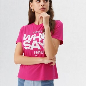 May include: A magenta t-shirt with white text that reads "WHO SAY" in a bold, stylized font. Below the English text is text in a different language. The model is wearing blue jeans.