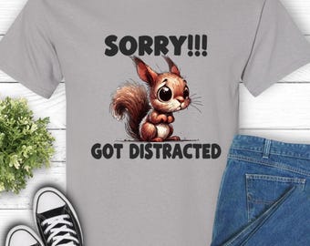 May include: A light gray t-shirt featuring a cartoon squirrel with large eyes. The squirrel is brown and white, with the text "SORRY!!! GOT DISTRACTED" in black. The shirt is paired with blue jeans and black sneakers.