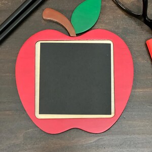 May include: A red apple-shaped chalkboard with a black writing surface, a light wood border, and a green leaf and brown stem. Two pencils and a pair of glasses are in the background. A teacher's desk accessory.