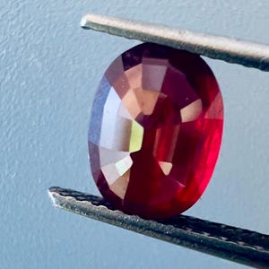 Pode incluir: Uma gema de rubi oval e facetada, segurada por uma pinça. A gema exibe uma cor vermelha profunda com reflexos internos. A pinça é prateada e o fundo é azul claro.