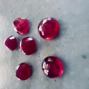 May include: A collection of faceted, deep red gemstones. The gems vary in size and shape, with a brilliant, reflective surface. The stones are scattered on a light gray surface, showcasing their rich color and intricate cuts.