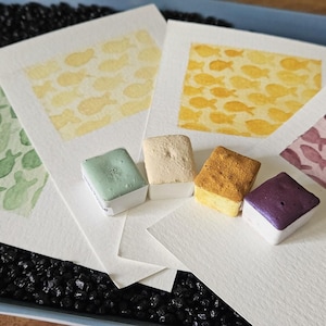 Peut inclure: Quatre feuilles de papier aquarelle rectangulaires présentant des motifs de poissons répétitifs en vert clair, jaune pâle, jaune doré et violet sourd, accompagnées de quatre godets d'aquarelle carrés assortis.
