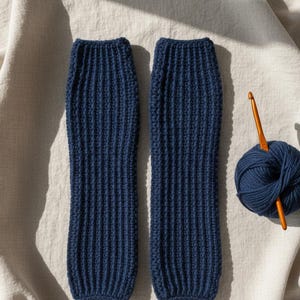 Op de afbeelding: Twee marineblauwe gebreide beenwarmers met een geribbelde textuur worden getoond naast een bol wol en een haaknaald. De beenwarmers zijn gemaakt van een zacht, getextureerd materiaal. Garen en haaknaald suggereren handgemaakt.