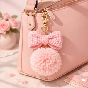 Può includere: Un portachiavi rosa con fiocco e pompon all'uncinetto appeso a una borsa rosa. Il fiocco è realizzato con un filato strutturato e il pompon è soffice. Il portachiavi ha una chiusura color oro.