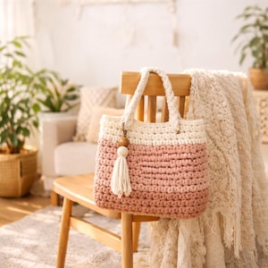 Może przedstawiać: Ręcznie robiona torba typu tote z szydełka w kolorze kremowym i pudrowym różu, z kremową rączką i frędzlem. Torba wisi na drewnianym krześle. Torba ma drewniany koralik i urok frędzla.