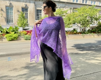 May include: A woman wearing a purple lace shawl over a black maxi dress. She is standing on a sidewalk in front of a building.