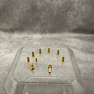 May include: A collection of small, gold-colored metal rivets and eyelets of varying sizes. The rivets have a cylindrical shape with a small hole in the center. They are displayed on a clear, angled surface.