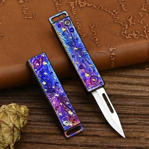 Könnte beinhalten: Zwei kleine, dekorative Klappmesser. Die Griffe sind eine Mischung aus Blau, Lila und Gold mit einem feuerwerksartigen Design. Die Klingen sind silberfarben. Die Messer liegen auf einer Holzoberfläche.