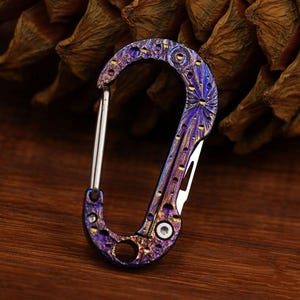 May include: A multi-coloured carabiner with an integrated knife. The carabiner is mainly purple and blue with gold accents and a silver clasp. The knife blade is silver. The carabiner is on a wooden surface.
